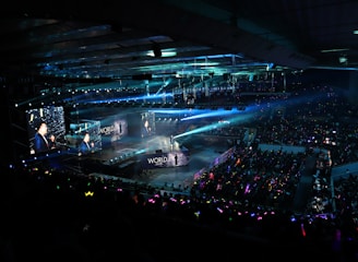 A large indoor arena filled with an audience watching a live performance. The stage features large screens displaying a performer and graphics, with bright spotlights and ambient lighting creating a dynamic atmosphere. The crowd is holding glow sticks, adding vibrant colors to the scene.