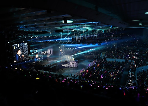 A large indoor arena filled with an audience watching a live performance. The stage features large screens displaying a performer and graphics, with bright spotlights and ambient lighting creating a dynamic atmosphere. The crowd is holding glow sticks, adding vibrant colors to the scene.