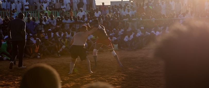 A vibrant wrestling match taking place outdoors, surrounded by a large crowd of spectators. The atmosphere is charged with excitement as people stand and sit around the dirt arena, intently watching the competitors. The sun is setting, casting a warm glow over the scene.