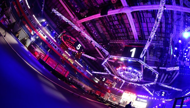 An indoor arena with vibrant purple and blue lighting, featuring large numbered screens suspended from the ceiling. The venue has a high ceiling with intricate metal truss structures and hanging lights, creating a futuristic and energetic atmosphere.