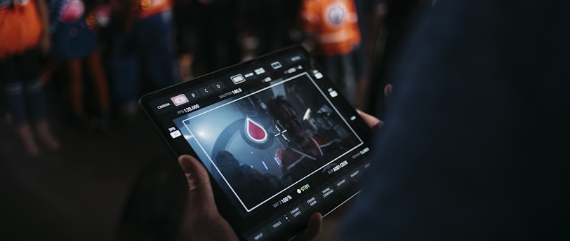 A person is holding a tablet that displays a video or image of a red and blue emblem on the screen. In the background, several people wearing dark and orange colored clothing, possibly sports jerseys, are partially visible. The scene is dimly lit, suggesting an indoor or evening setting.