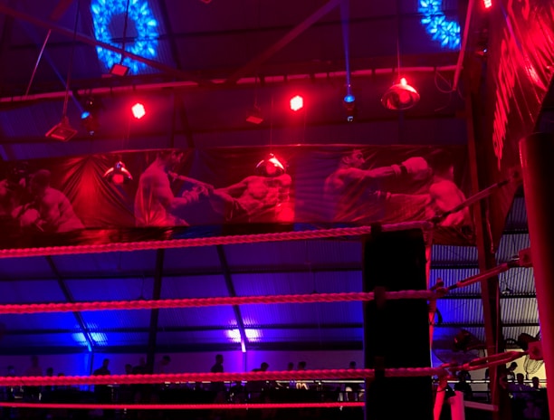 A brightly lit boxing or wrestling ring with vivid red and blue lights. Banners with images of fighters hang from the ceiling. The ropes of the ring are prominently visible, and a stool is placed inside the ring. The arena is filled with an audience, silhouetted against the colorful lighting.