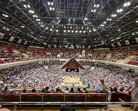 A large indoor arena packed with spectators seated in tiers around a central platform. The platform features traditional Japanese decor, indicating it may be used for sumo wrestling. The atmosphere is bustling, with people filling the seats and many spectators visible in the foreground.