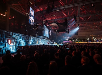 A large indoor event with a stage featuring bright screens and spotlights, attended by a substantial crowd. The venue has a high ceiling with industrial beams and red lighting accents. The atmosphere resembles a concert or a conference with a focus on visual effects.